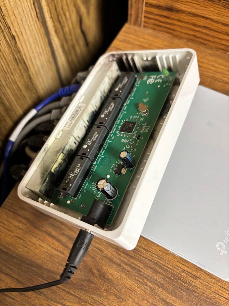 Open white plastic box showing internal circuitry with green circuit board, black battery compartment, and connected blue and black cables on wooden desk